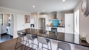 A modern kitchen with a central island, bar stools, a built-in oven, and a stainless steel range hood. - SeaRenity - Waimarama Holiday Home (Havelock North)
