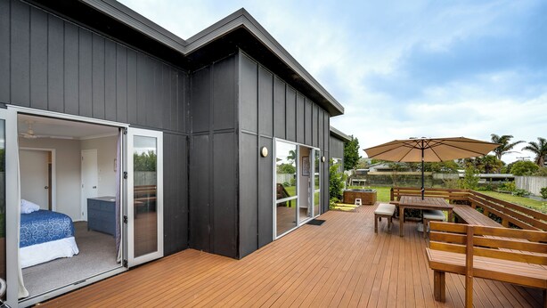 A modern house with a deck, a bedroom with a bed and dresser, and a patio with a table and chairs. - SeaRenity - Waimarama Holiday Home (Havelock North)
