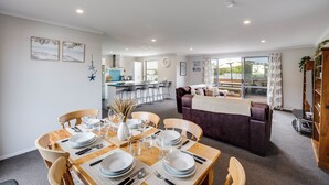 A dining area with a wooden table set for four, a sofa, and a kitchen in the background. - SeaRenity - Waimarama Holiday Home (Havelock North)