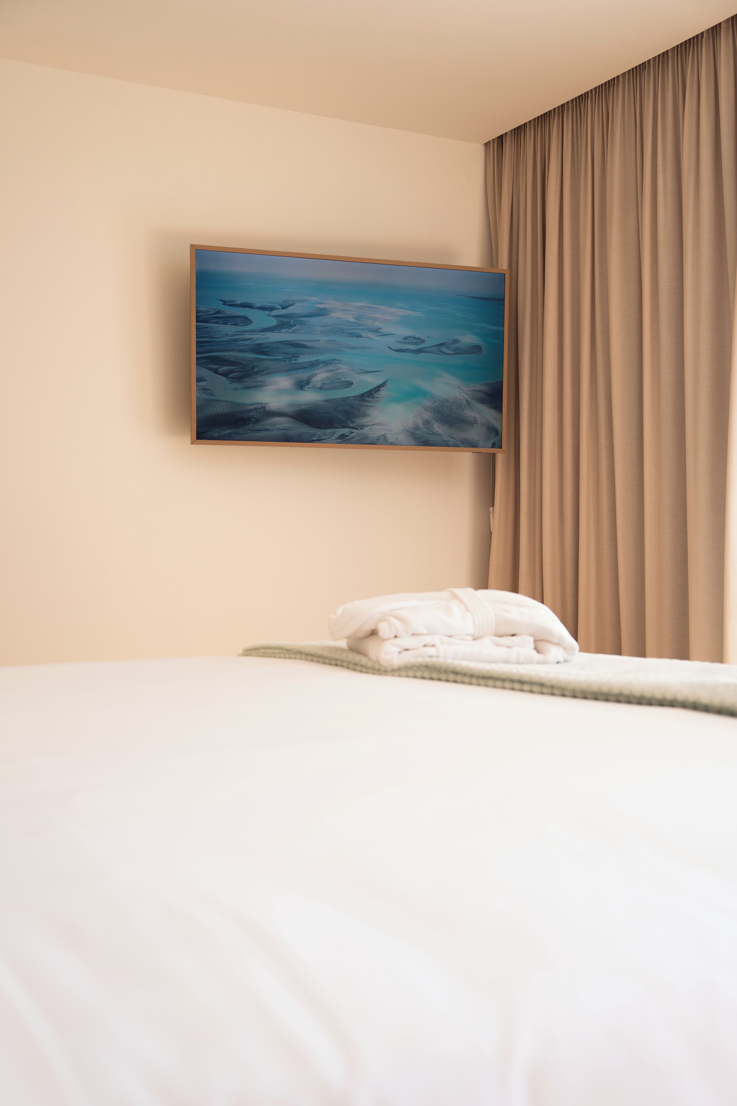 Une chambre d’hôtel avec un lit, une serviette blanche soigneusement pliée, un tableau représentant un paysage marin et des rideaux de couleur beige.