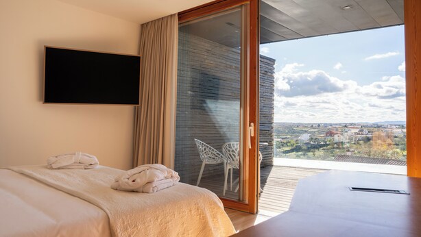 A modern bedroom with a bed, a TV mounted on the wall, and a sliding glass door leading to a balcony with a view of the countryside. - Foz Côa Story House (Vila Nova de Foz Côa)