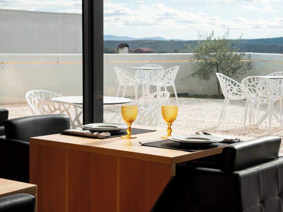 A dining area with a wooden table and chairs, two golden goblets, and a view of the landscape.