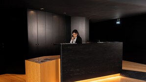 A reception desk with a woman standing behind it, a wooden counter, and a dark wall with a ceiling light. - Foz Côa Story House (Vila Nova de Foz Côa)