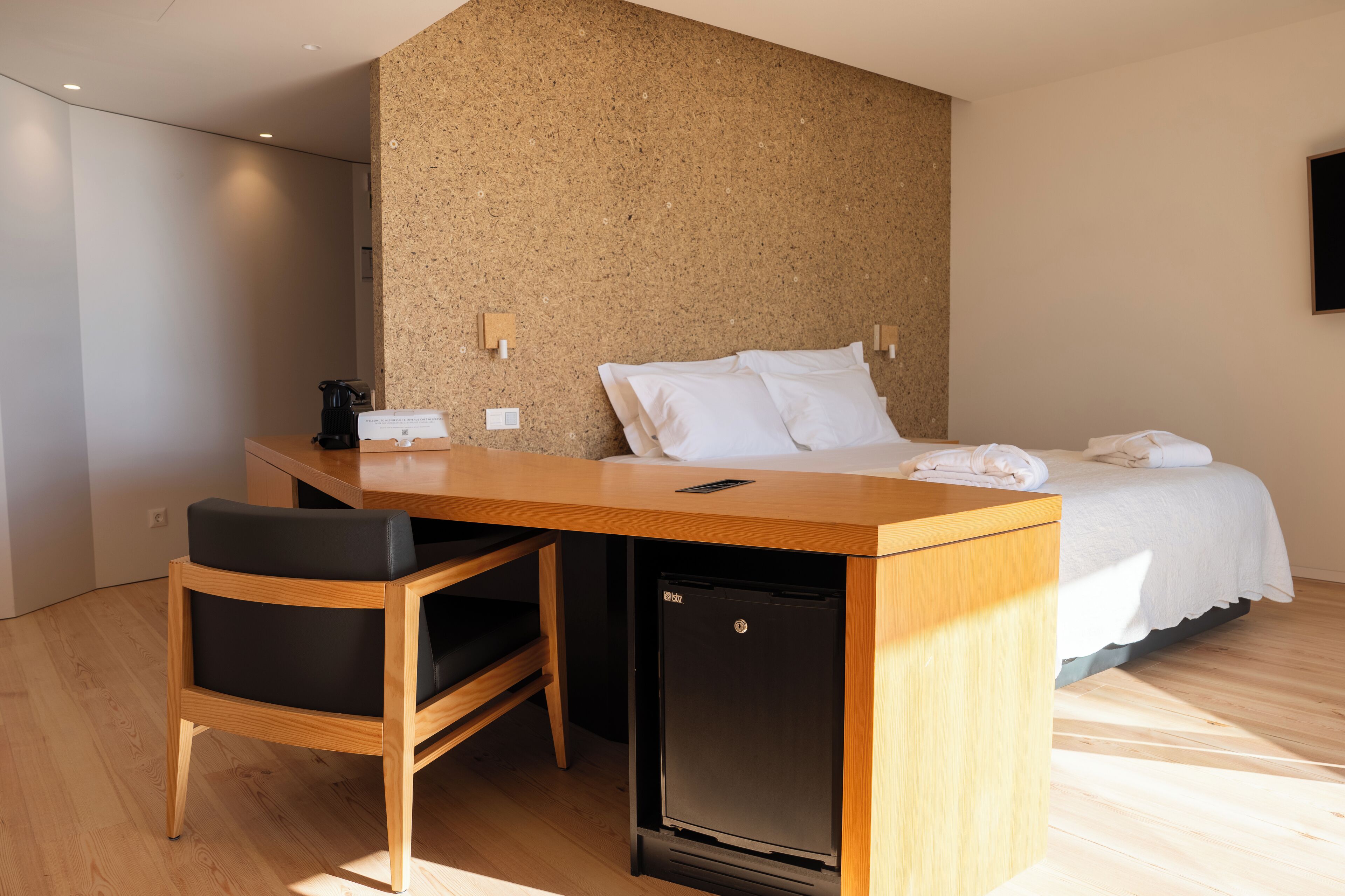 A hotel room with a wooden desk, a black chair, a bed with white linens, and a small black refrigerator.