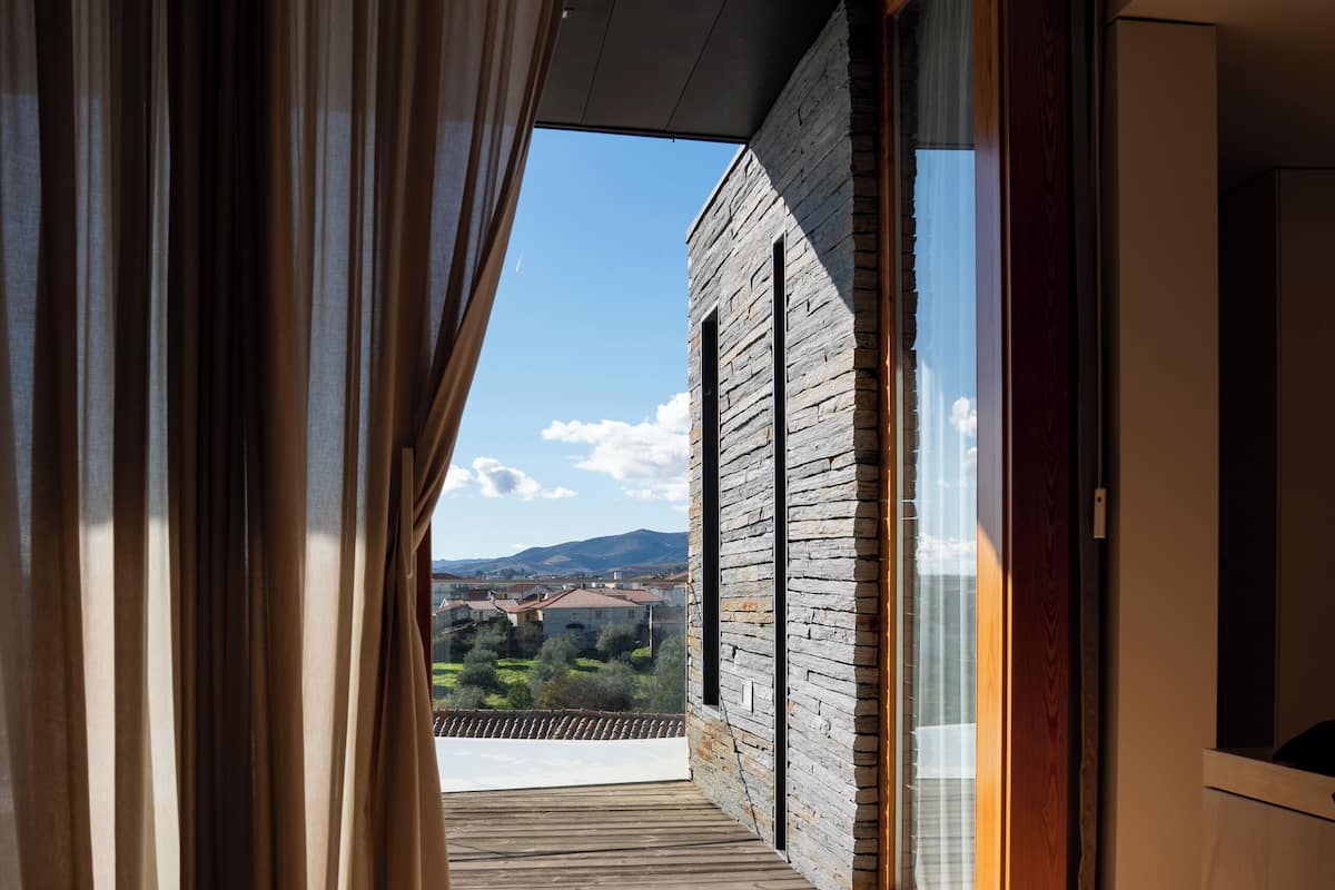 Un dormitorio con suelo de madera, una cama y vistas a un balcón con pared de piedra y paisaje lejano.
