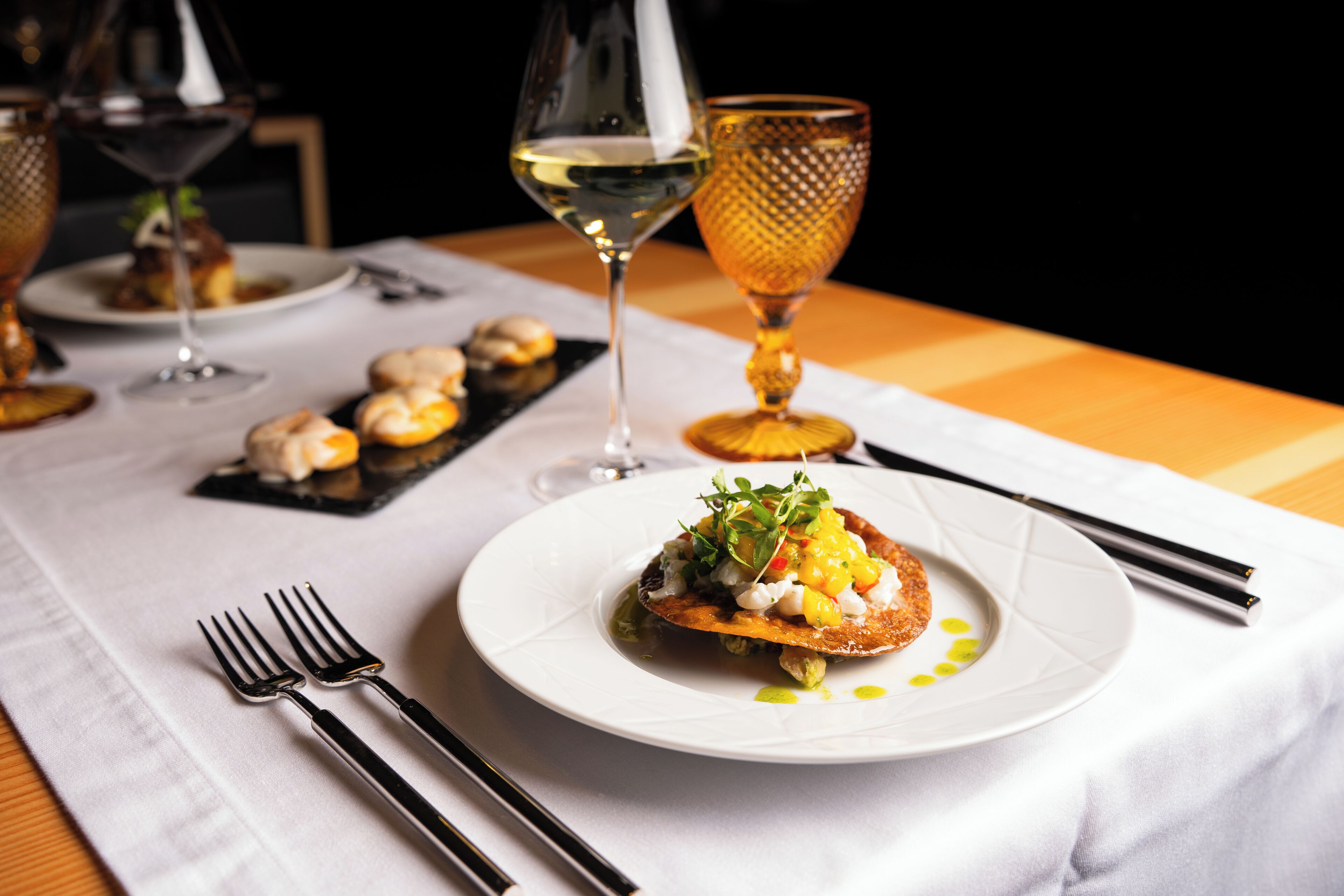 A dining table with a white tablecloth, two forks, a glass of white wine, and a plate of food.
