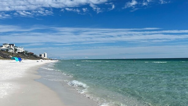 A beach with clear blue water, a sandy shore, and a few buildings in the background. - Private Beach + Chair Service/Pool/Bikes (Santa Rosa Beach)