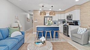 A modern kitchen with a central island, bar stools, and a dining area with a blue sofa and a grey armchair. - Private Beach + Chair Service/Pool/Bikes (Santa Rosa Beach)