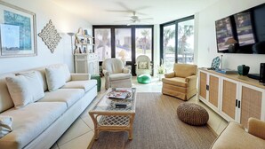 A modern living room with a white sofa, a glass coffee table, a wicker chair, a green ottoman, and a television on a cabinet. - Ground Floor Condo with Shared Pool, Golf Access, Bay Views, and WiFi (Panama City)