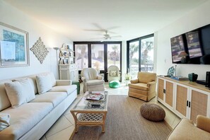 Ein modernes Wohnzimmer mit einem weißen Sofa, einem Glastisch, einem Korbstuhl, einem grünen Hocker und einem Fernseher auf einem Schrank.