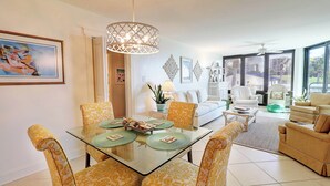 A modern living room with a glass dining table, yellow patterned chairs, a ceiling fan, and large windows offering a view of the outdoors. - Ground Floor Condo with Shared Pool, Golf Access, Bay Views, and WiFi (Panama City)