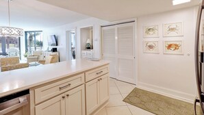 A modern kitchen with a large island, white cabinetry, and a dining area with a sofa and chairs. There are framed pictures on the wall and a door with louvered shutters. - Ground Floor Condo with Shared Pool, Golf Access, Bay Views, and WiFi (Panama City)