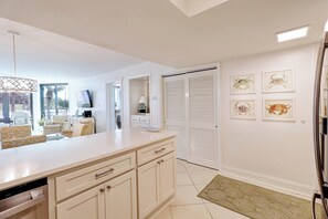Eine moderne Küche mit einer großen Kochinsel, weißen Schränken und einem Essbereich mit einer Couch und Stühlen. An der Wand befinden sich gerahmte Bilder und eine Tür mit Klappläden.