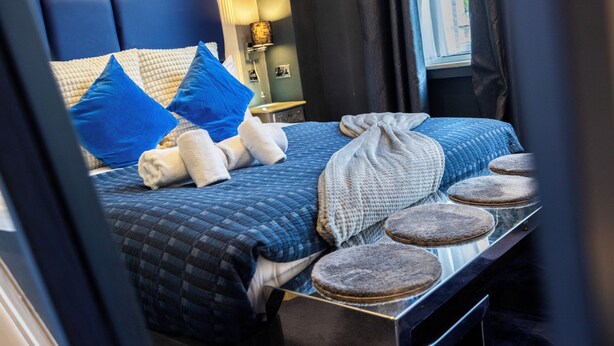 A bed with blue and white bedding, a bedside table with a lamp, and a window with curtains. - Stunning Super king & Double Bed Apartment|Central London|Oxford St|Bond St (London)