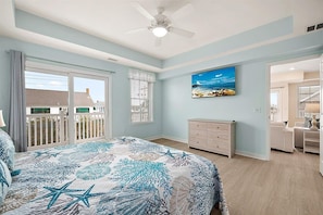 A bedroom with a bed, a dresser, a ceiling fan, a painting, and a view of the outside. - Oceanside Condo Steps from the Beach and Boardwalk (Ocean City)