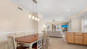 Angolo ristoro con tavolo in legno e sedie bianche, cucina con mobili in legno e vista sulla spiaggia attraverso ampie finestre.