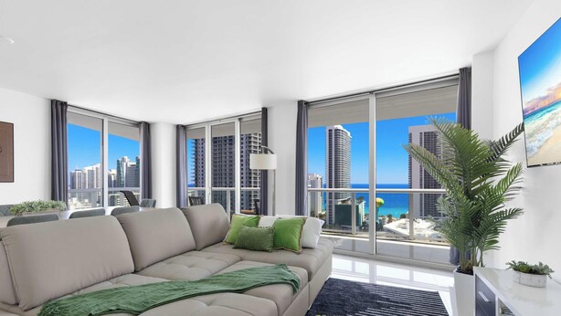 Ein modernes Wohnzimmer mit einer großen Couch, einem Couchtisch und Blick auf Stadt und Strand durch raumhohe Fenster.