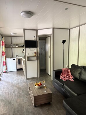 A modern living room with a black leather sofa, a wooden coffee table with fruit and a glass of wine, a TV mounted on the wall, and a kitchen area visible through an open door. - Magnificent 3-bedroom chalet. Ideal spot for kitesurfing and lounging! (Le Vauclin)