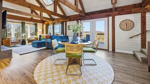 A spacious living area with a high ceiling, wooden beams, a round table with chairs, and a large window providing a view of the outdoors. - Mtn Views | Stargazing Dome | Hot Tub | Sleeps 20 (Fairview)