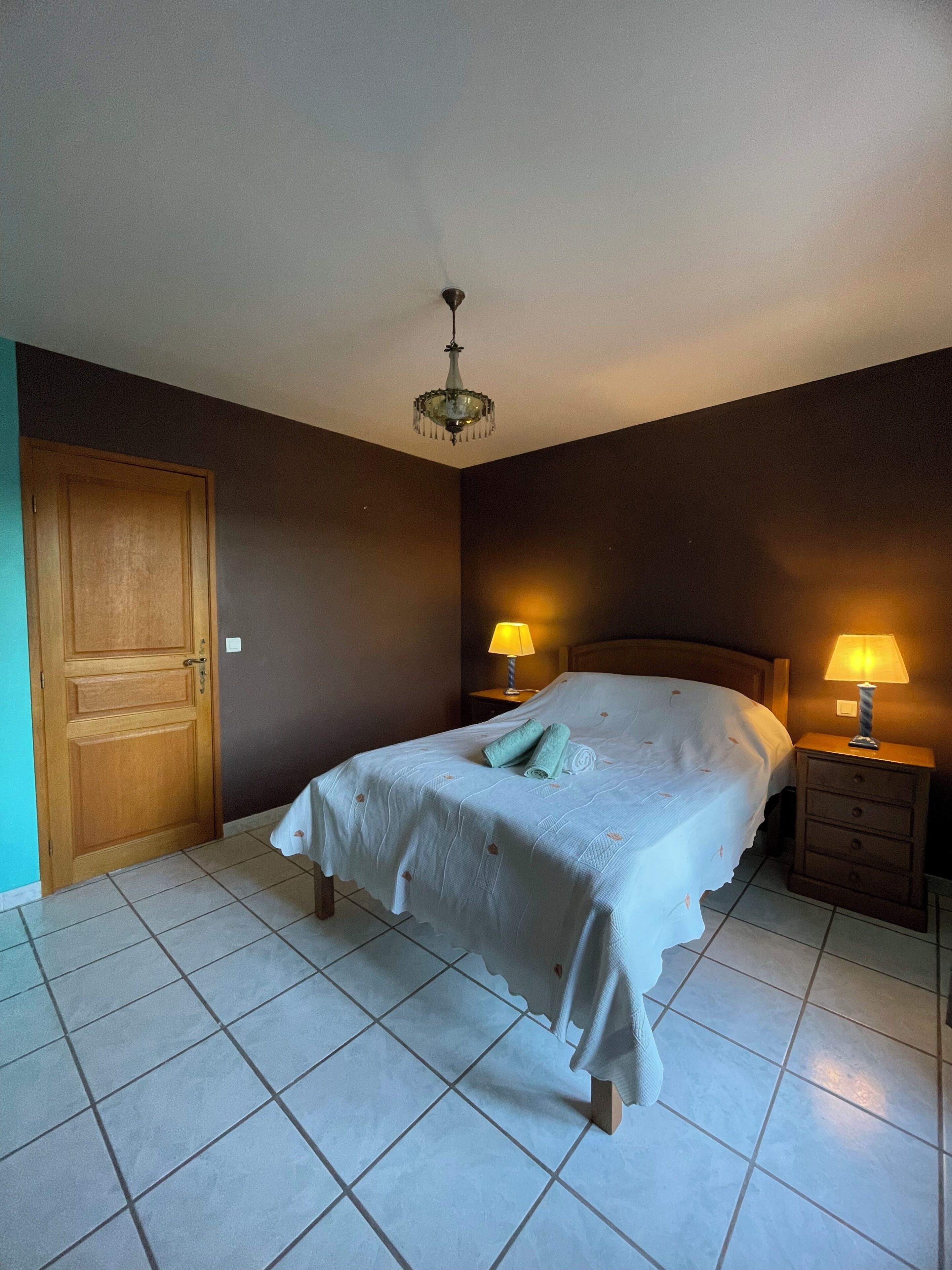 Ein Schlafzimmer mit einem Bett, zwei Nachttischlampen, einer Kronleuchter, einer Holztür und gefliestem Boden.