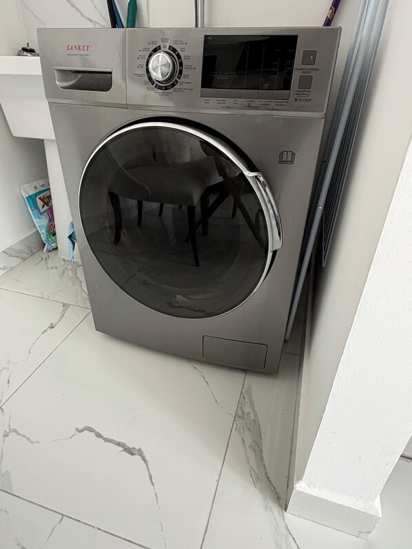 Un appareil combiné lave-linge et sèche-linge à chargement frontal, en acier inoxydable, doté d’un panneau de commande, installé dans une salle de bain carrelée de marbre blanc.