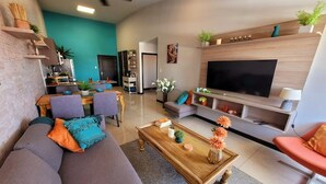 A modern living room with a sofa, coffee table, and TV. There is a dining area with chairs and a table. The room has a teal accent wall and wooden shelves. - Near Airport, Lush Gardens, Pool, King Bed, Security SANTA ANA NEAR DOWNTOWN (Santa Ana)