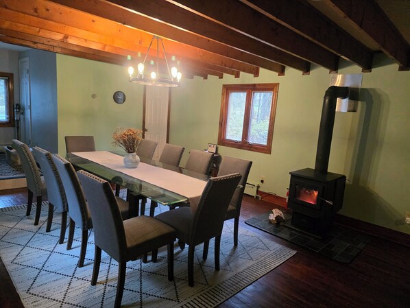 A dining area with a wooden ceiling, a chandelier, a table with chairs, a vase with flowers, a window, and a wood-burning stove.