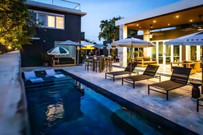 A modern outdoor pool area with lounge chairs and umbrellas, surrounded by a well-lit patio and a building with large windows. - Portofino 4BD Suite | Resort Vibe Near Beach | Pool (San Juan)