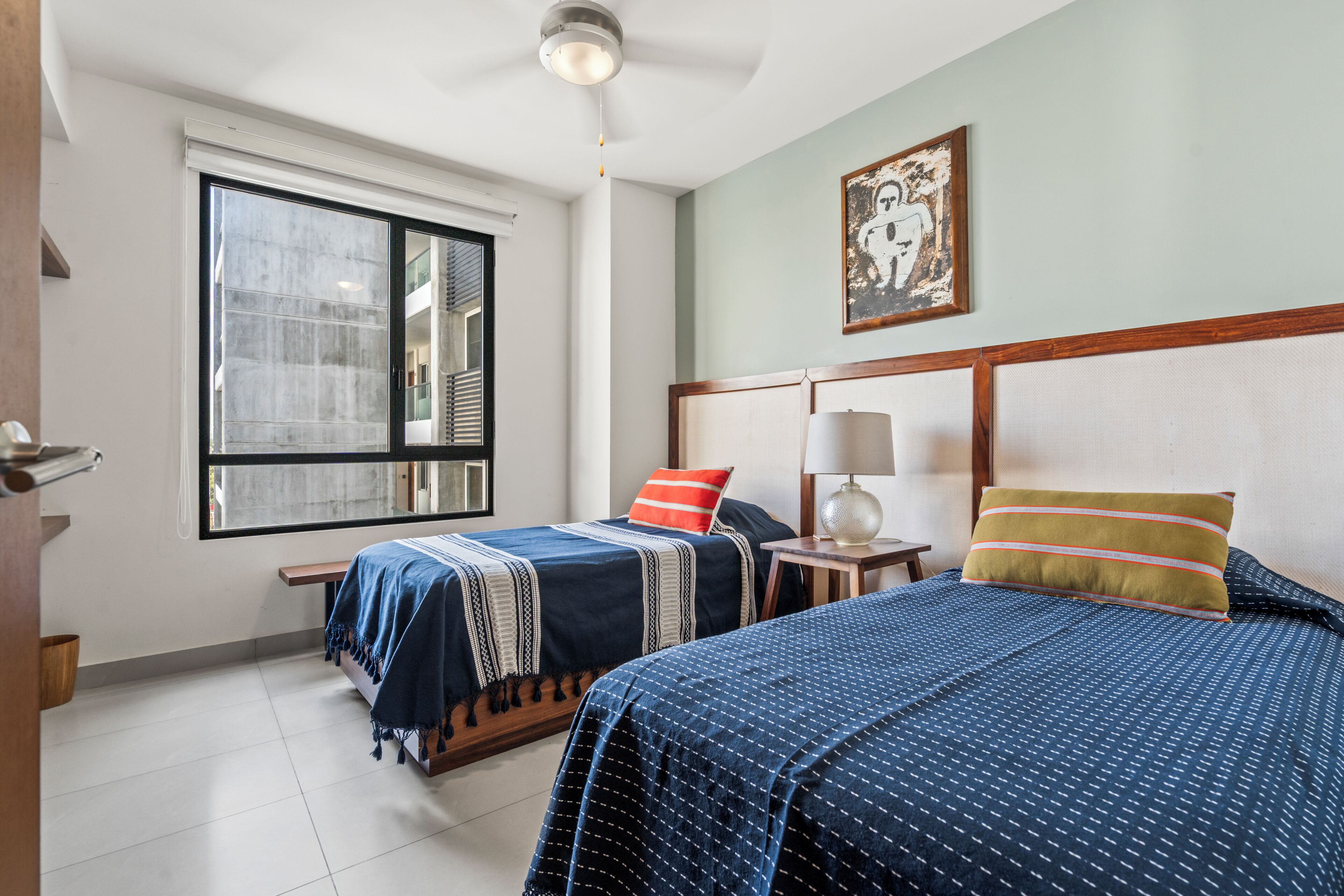 A bedroom with two beds, a window, a ceiling fan, and a painting on the wall.