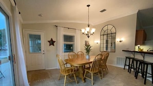 A dining area with a round wooden table and six chairs, a chandelier, and a kitchen area with stools. - Pet friendly Charming Bonnybrook Villa! (The Villages)