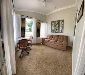 A room with a beige carpet, a brown sofa, a round table with a chair, and a window with white curtains. - Pet-Friendly Villa Near Lake Sumter & Pool (The Villages)