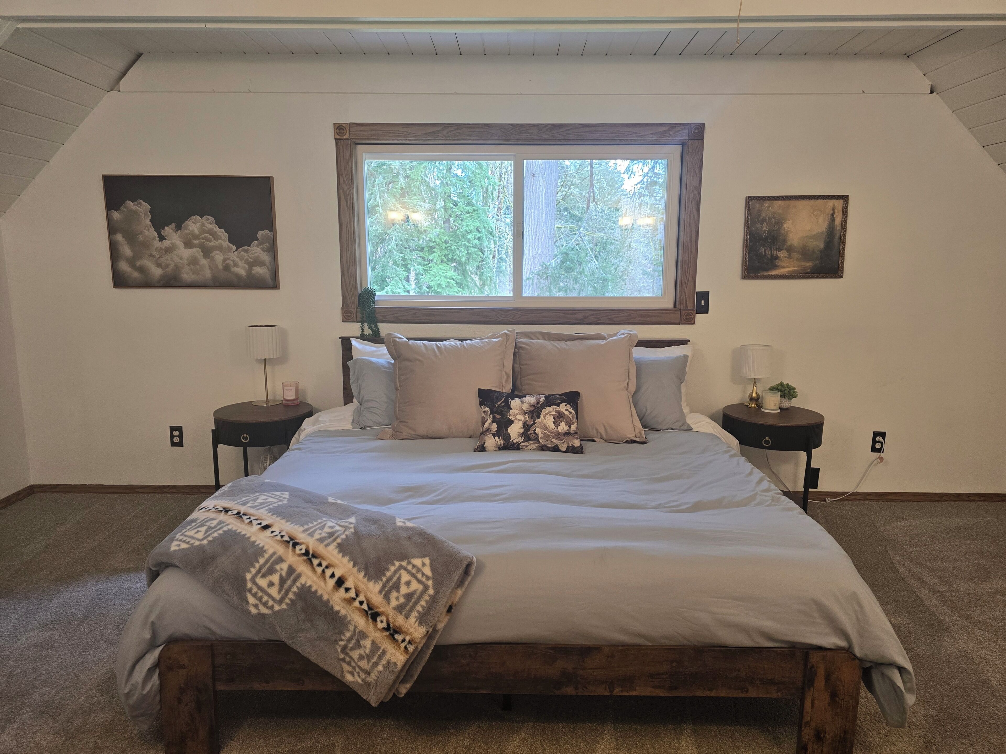 Un dormitorio con una cama grande, dos mesitas de noche, un ventanal con vistas a árboles y dos obras de arte colgadas en la pared.