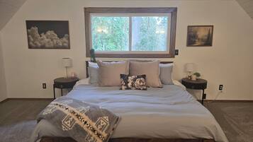 Un dormitorio con una cama grande, dos mesitas de noche, un ventanal con vistas a árboles y dos obras de arte colgadas en la pared.