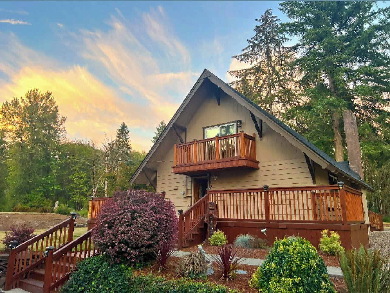 A two-story log cabin with a balcony, surrounded by a landscaped garden with various shrubs and trees.
