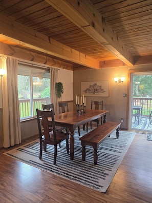 A wooden dining room with a large table, chairs, and a bench. There is a window with curtains, a wall-mounted light fixture, and a framed picture. - Peaceful Cabin Retreat Near Mayfield Lake- Boating, Fishing, Hunting, Hiking (Silver Creek)