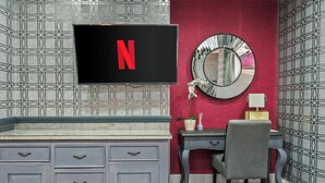 Een kamer met een televisie waarop het Netflix-logo staat, een bureau met een stoel, een spiegel en behang met een patroon.