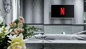 Een moderne kamer met een grijze bank, een aan de muur bevestigde televisie met daarop het Netflix logo, en een klok.