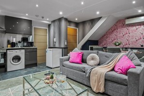 Un espacio de estar moderno con sofá gris, una mesa de centro de cristal, lavadora y una zona de cocina con microondas.