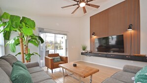 Ein modernes Wohnzimmer mit einer großen Couch, einem Couchtisch, einem an einer Holzvertäfelung befestigten Fernseher und einem Deckenventilator.