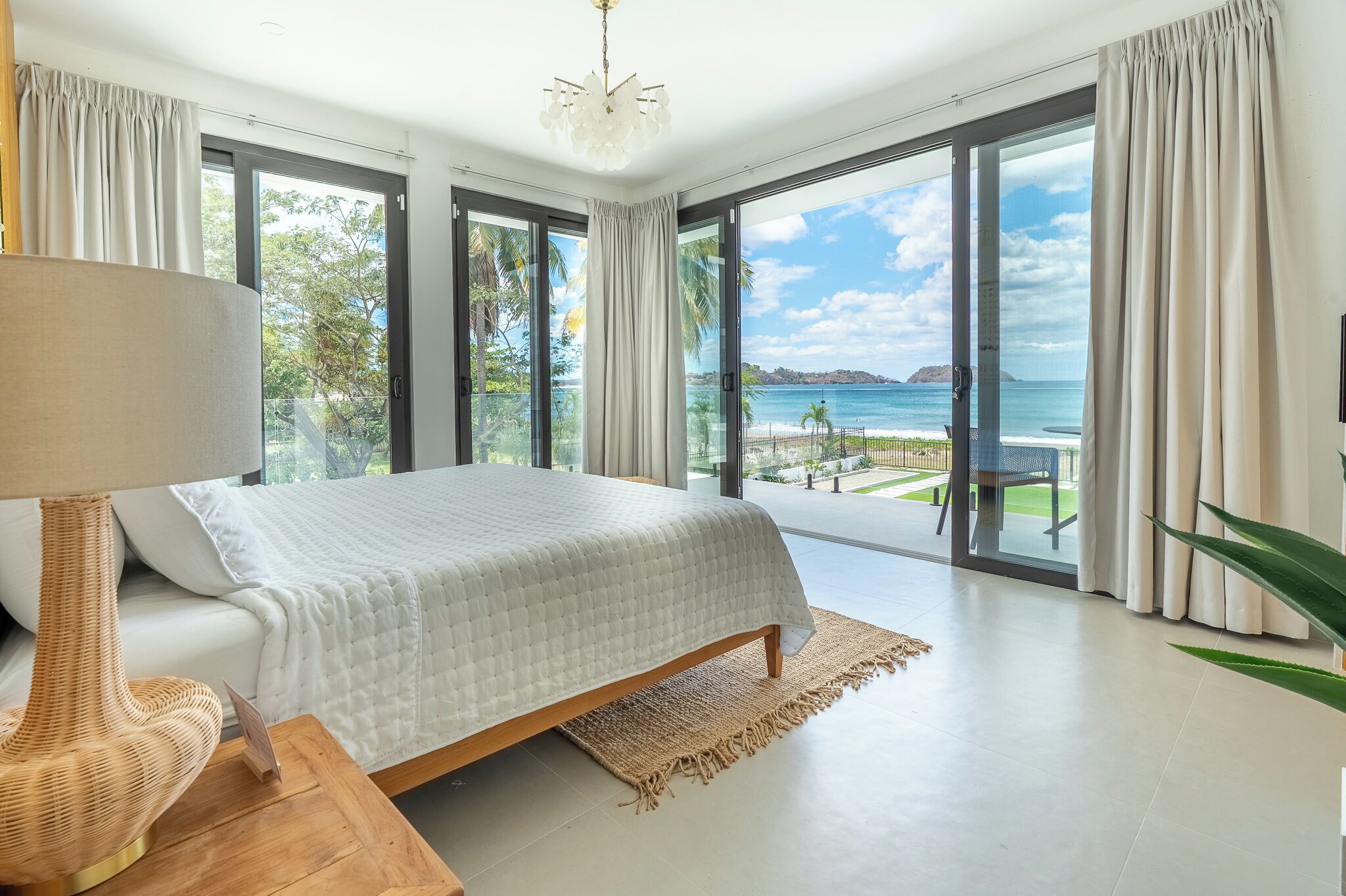 A bedroom with a large bed, a bedside lamp, and a view of the ocean through floor-to-ceiling windows.