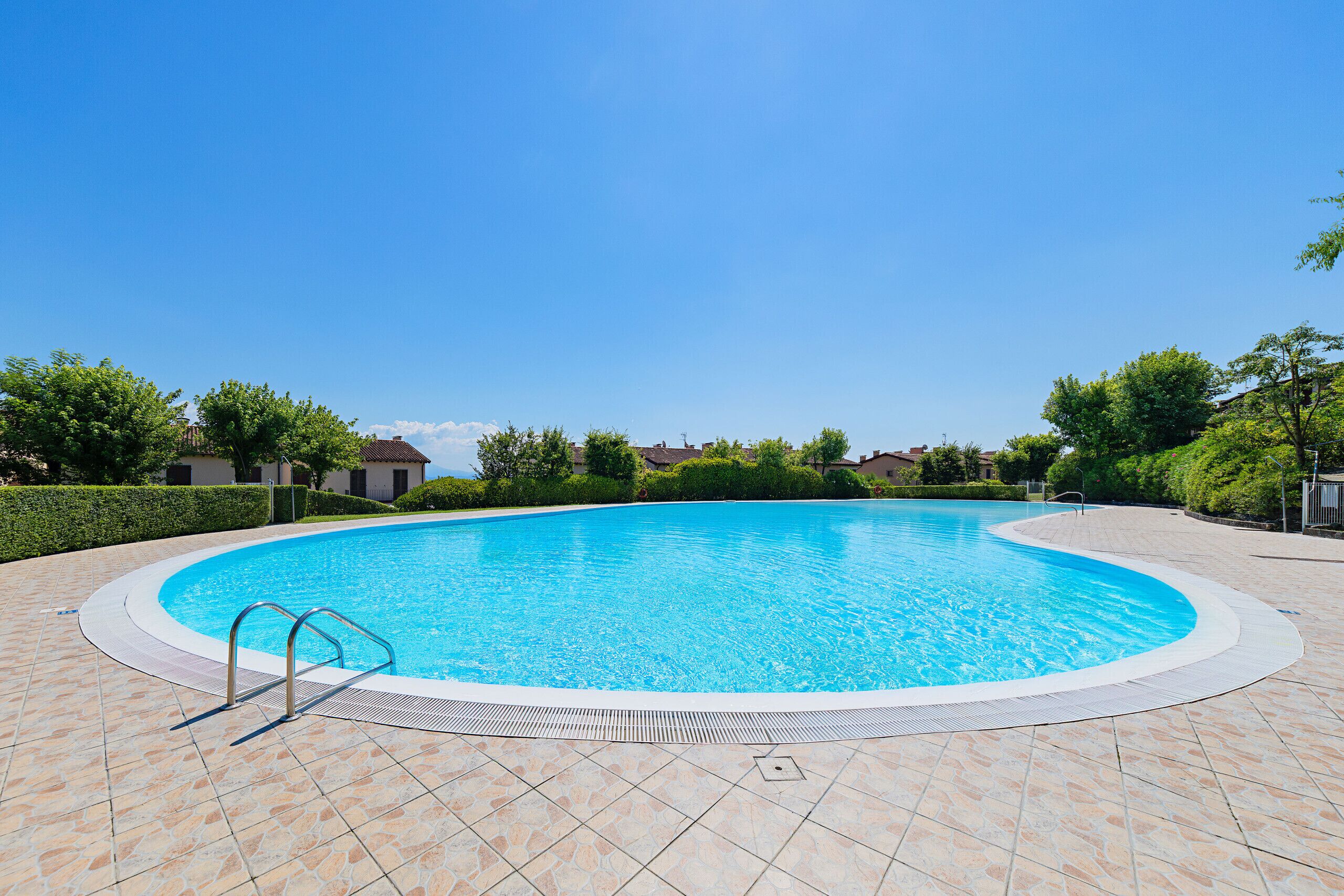 A clear blue swimming pool with a tiled deck, surrounded by greenery and residential buildings.