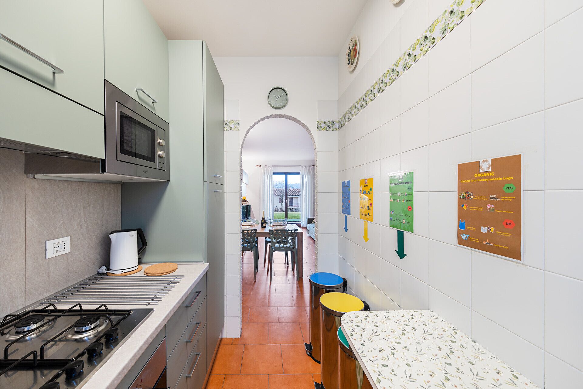 A narrow kitchen with a gas stove, microwave, and a dining area with a table and chairs. There are recycling and organic waste disposal signs on the wall.