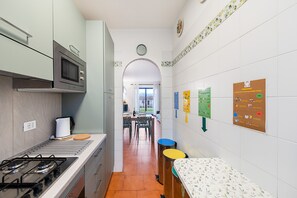 Una cucina piccola con piano cottura a gas, forno a microonde e zona pranzo con tavolo e sedie. Sulle pareti, cartelli per la raccolta differenziata e lo smaltimento dei rifiuti organici.