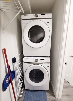 A laundry room with two stacked washing machines, a drying rack, and cleaning tools. - Newly Furnished Private Suite | Separate Entry, Kitchen, Laundry & Workspace (Regina)