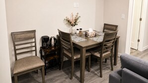 Un comedor con una mesa de madera, cuatro sillas, una cafetera, un florero con flores y una caja de pañuelos.
