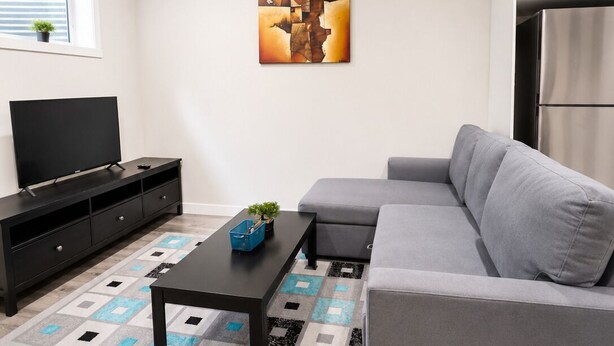 Un salón moderno con un sofá gris, una mesa de centro negra, un televisor de pantalla plana sobre un soporte negro y un cuadro en la pared.