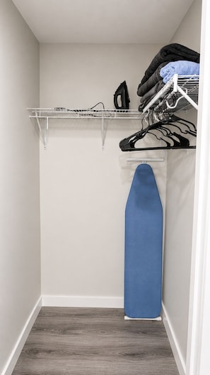 A closet with a hanging rack, an ironing board, and a shelf with folded clothes.
