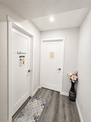 A hallway with two white doors, one labeled 'bathroom' and the other with a poster. A patterned rug leads to the doors, and a vase with flowers is placed on the floor to the right. - Newly Furnished Private Suite | Separate Entry, Kitchen, Laundry & Workspace (Regina)