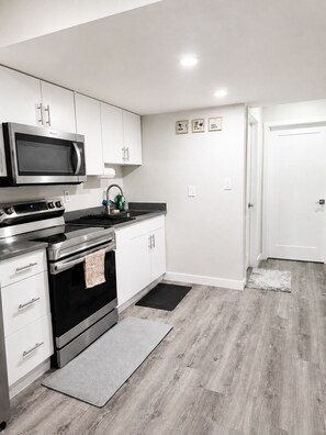 A modern kitchen with white cabinetry, a stainless steel stove, a microwave, and a sink. There are three framed pictures on the wall and a door to the right. - Newly Furnished Private Suite | Separate Entry, Kitchen, Laundry & Workspace (Regina)