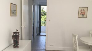 Ein Zimmer mit einem weißen Schrank, einem kleinen Tisch mit einem Buch und einem Stuhl. Eine Tür führt zu einem Balkon mit Blick auf Grünflächen.